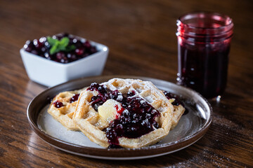 Close-up of golden waffles topped with butter, fresh huckleberries, and berry jam as powdered sugar is sprinkled on top. Rustic breakfast or dessert food photography with a gourmet, homemade style.