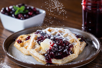 Close-up of golden waffles topped with butter, fresh huckleberries, and berry jam as powdered sugar is sprinkled on top. Rustic breakfast or dessert food photography with a gourmet, homemade style.