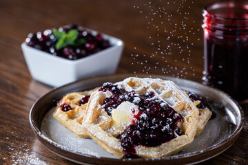 Close-up of golden waffles topped with butter, fresh huckleberries, and berry jam as powdered sugar is sprinkled on top. Rustic breakfast or dessert food photography with a gourmet, homemade style.