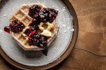Close-up of golden waffles topped with butter, fresh huckleberries, and berry jam as powdered sugar is sprinkled on top. Rustic breakfast or dessert food photography with a gourmet, homemade style.