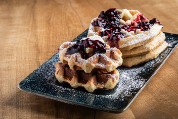 Close-up of golden waffles topped with butter, fresh huckleberries, and berry jam as powdered sugar is sprinkled on top. Rustic breakfast or dessert food photography with a gourmet, homemade style.