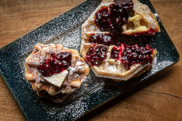 Close-up of golden waffles topped with butter, fresh huckleberries, and berry jam as powdered sugar is sprinkled on top. Rustic breakfast or dessert food photography with a gourmet, homemade style.