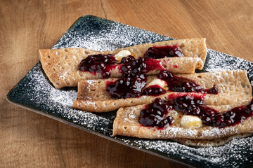 Close-up of homemade crepes topped with fresh huckleberries, berry jam, and powdered sugar on a rustic wooden table.