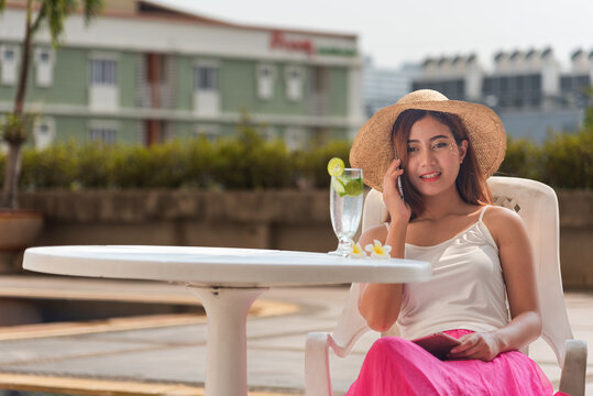 Asian women drinking lime juice in blue lagoon swimming pool summer time. Happy female tropical drink with summer dress, straw hat at hotel smile face at pool outdoors. Young traveller poolside