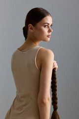 Young woman with long braided hair wearing a beige sleeveless top stands thoughtfully against a...