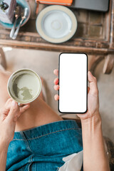 Overhead view of a woman holding a coffee cup and smartphone with a blank screen, ready for customization.
