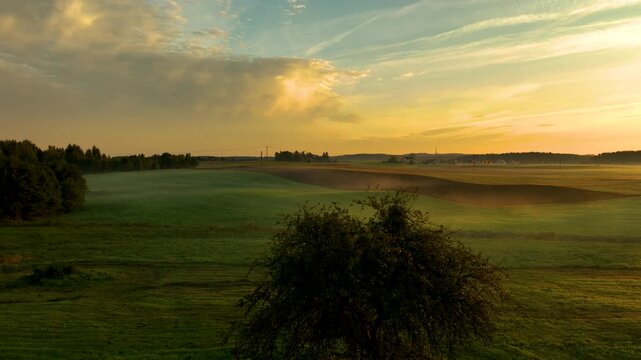 Golden sunlight breaks through wispy clouds over a misty, rolling green field. A wide, atmospheric aerial view of a peaceful, rural sunrise