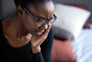 An African American woman in glasses shows signs of distress while sitting in a comfortable room filled with cushions. Her expression reveals depression and deep emotional concern. Generative AI.