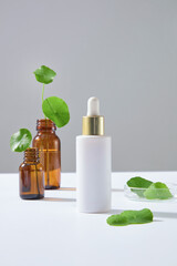 White cosmetic bottle displayed with Centella leaves and amber dropper bottles. Clean minimalist look for natural ingredient-based skincare advertising. Serum bottle mockup design