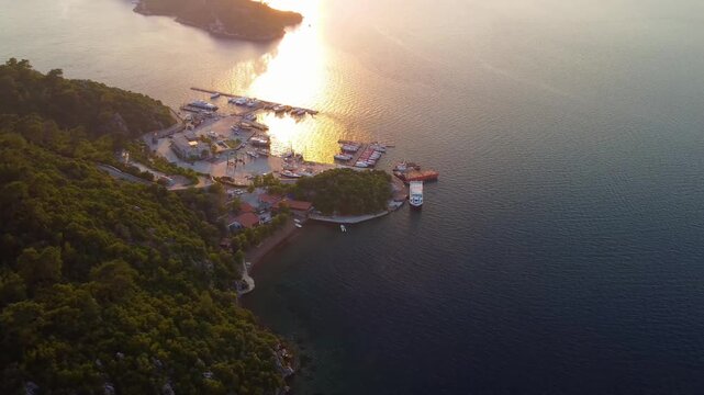 aerial perspective captures the golden hour over Cennet Adasi Paradise Island, a stunning peninsula near Marmaris, Turkey. The footage showcases the serene marina filled with yachts, surrounded by lus