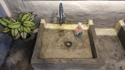 Old concrete sink with a rusty drain, faucet, and a hand soap bottle on top. A green ornamental plant decorates the side, giving contrast between hygiene, rustic interior, and poor maintenance. Suitab