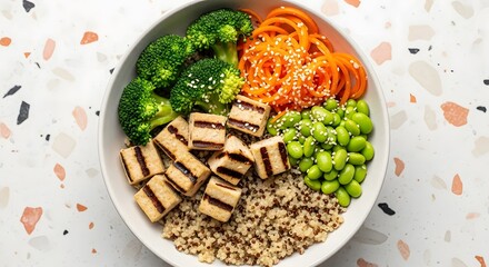 A vibrant and healthy vegan buddha bowl filled with grilled tofu, quinoa, fresh broccoli, edamame, and spiralized carrots, presented from a top-down perspective on a terrazzo surface