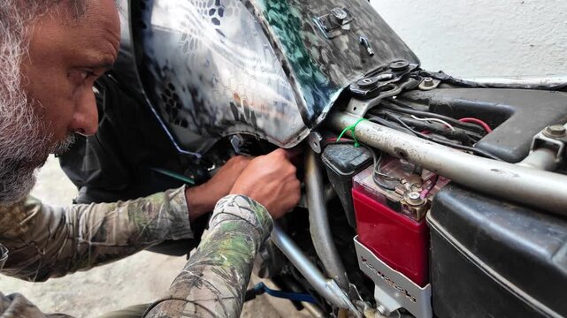 Focused mechanic troubleshooting the electrical system and ignition relay of a camouflage adventure motorcycle in a home workshop.