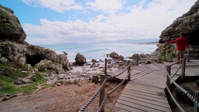 Male traveler on wooden boardwalk at historic Klipgat Cave looks out over ocean