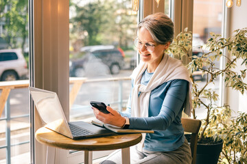 Smiling senior woman checking smartphone while working in a cafe with laptop and notebook