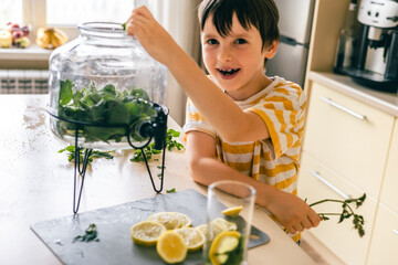 Smiling boy adding fresh mint to jar for lemonade, making summer drink at home in kitchen