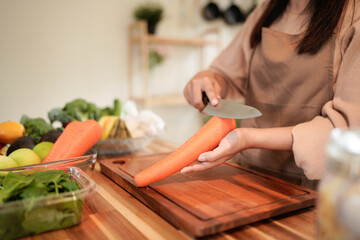 Obraz premium Asian young woman, girl or housewife hand using knife, cutting carrots on board, on wooden table in kitchen home, preparing ingredient, recipe fresh vegetables for cooking meal. Healthy food people.