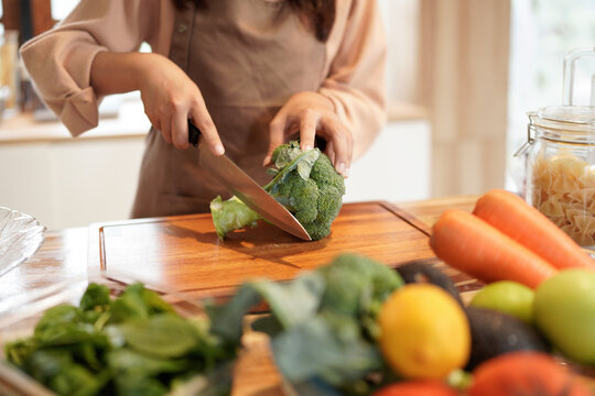 A woman is cutting a broccoli on a wooden cutting board. The kitchen is well-stocked with various fruits and vegetables, including apples, carrots