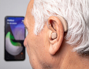 AI generated elderly person ear with a nearly invisible hearing aid, displaying a smartphone soundscape map filtering specific sounds in a busy environment. Focus on technology and discretion.