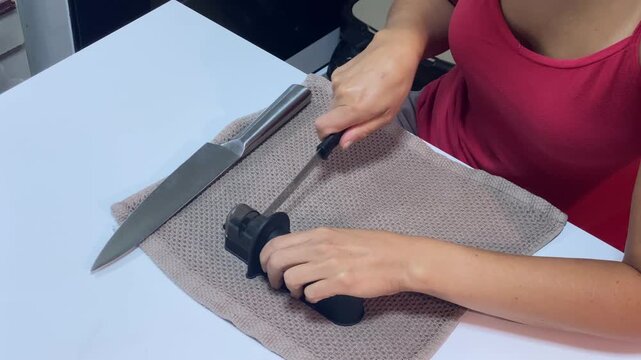 A person's hands are actively using a knife sharpener on a kitchen knife, placed on a towel on a white table. This action prepares the tool for cooking or other precise tasks.