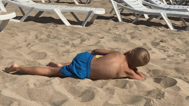 A young boy in blue swim trunks relaxes on a sandy beach, lying prone with a yellow shovel nearby, observing his surroundings on a bright day.