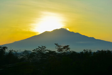 Dramatic sunrise over Mount Merbabu in Central Java, Indonesia, with the golden morning light creating a stunning silhouette of the mountain and the surrounding trees. The scene radiates beauty