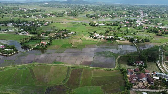 Drone video captures lush green fields and scattered houses under clear skies. The expansive landscape offers a serene rural vista with vibrant colors and intricate details.
