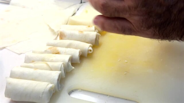 Hands are rolling thin pastry like yufka or phyllo to make Turkish cigarette pastry or spring rolls, with a row of prepared rolls on a white cutting board.