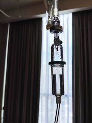 A low-angle shot captures an intravenous (IV) infusion tube about to drip, against the backdrop of a window in a somewhat dark hospital room.