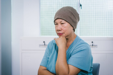 An elderly Asian woman wearing a gray-brown beanie, a light blue shirt, and dark pants sits quietly...