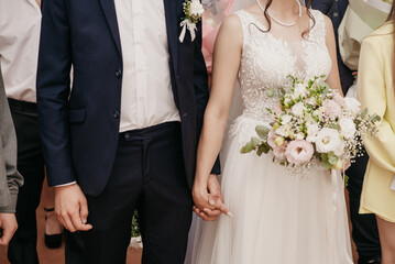 Groom in black suit holds bride in white dress embroidered with rhinestones by the hand while she holds bouquet of white roses during wedding ceremony