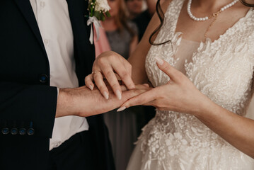 The bride in a white dress of modern design puts a gold wedding ring on the finger of the groom in a black classic suit during the wedding ceremony