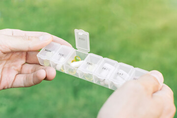 Weekly pill organizer with compartments for each day of the week filled with capsules and tablets held in hands against green outdoor background
