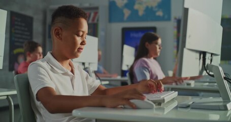 African American Schoolboy in White Polo Shirt is Concentrating on Work at Computer in Modern Classroom. In Background, Children Also Studying at Monitors. Digital Education, Technology. Handheld. - Powered by Adobe