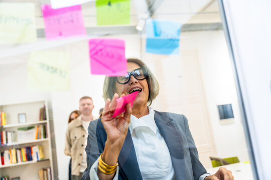 Mature businesswoman brainstorming ideas using sticky notes on glass - Powered by Adobe