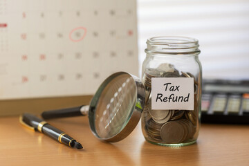 Tax refund planning concept. glass jar of coins labeled "Tax Refund" beside magnifying glass, pen, calendar deadline, and a calculator on desk. ideal for budgeting, filing season, and savings content.
