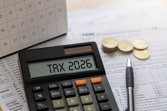 Annual tax planning concept with calculator displaying TAX 2026, stacks of coins, a calendar, and a pen on a wooden desk. ideal for filings, budgeting, business finance, deadlines, and savings tips.