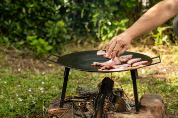 A person’s hand places raw bacon onto a cast iron griddle set over an open wood fire, in a rustic outdoor setting. The fire is built on bricks and surrounded by green grass and foliage, evoking a sens © Bojan