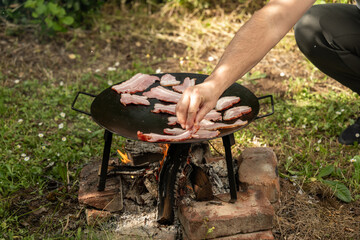 A person’s hand places raw bacon onto a cast iron griddle set over an open wood fire, in a rustic outdoor setting. The fire is built on bricks and surrounded by green grass and foliage, evoking a sens © Bojan