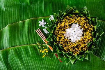 Krathong decorates with white and yellow flowers that have blurred focus of 3 incense sticks and candle put on banana leaf for Loy Krathong festival.