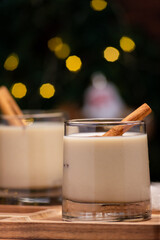 Two glasses of creamy eggnog with cinnamon sticks with festive holiday lighting in the background