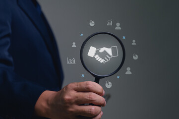 Businessman holds a magnifier over a handshake icon, symbolizing a deal or partnership. The concept is searching for agreement, collaboration, contract analysis, and building corporate trust.