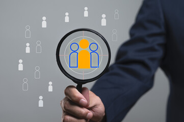 Businessman uses a magnifying glass to focus on a target candidate icon. A concept for human resources, recruitment, headhunting, and CRM strategy. Finding the right talent or customer for the team