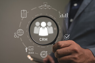 Businessman holds a magnifier over a CRM icon for Customer Relationship Management. A system for sales, marketing, and data analytics to manage customer interactions, retention, and business growth.