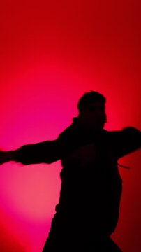 A man in a hoodie performs modern street dance as a clean silhouette against a vivid red gradient background in a minimalist studio, filmed in slow motion.