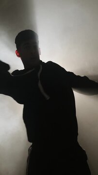 A man in a hoodie performs modern street dance against dense smoke with a strong beam backlight, creating a graphic silhouette in a minimalist studio, filmed in real time.