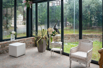 Elegant wedding ceremony setup inside a sunlit glass room overlooking a lush green garden with decorative floral arrangements and antique furniture perfect for intimate celebrations