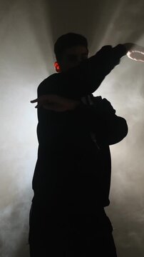 A man in a hoodie performs modern street dance against a smoky backlit background, creating a crisp silhouette in a minimalist studio, filmed in real time.