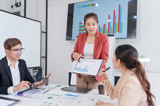 A dynamic business presentation scene where an Asian woman in a red blazer is presenting a healthcare business report to her colleagues.