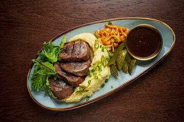 A plate of grilled meat, creamy mashed potatoes, pickles, and fresh greens
